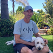 Man sitting on grass with a white dog in a sunny garden, wearing a cap and gray T-shirt.