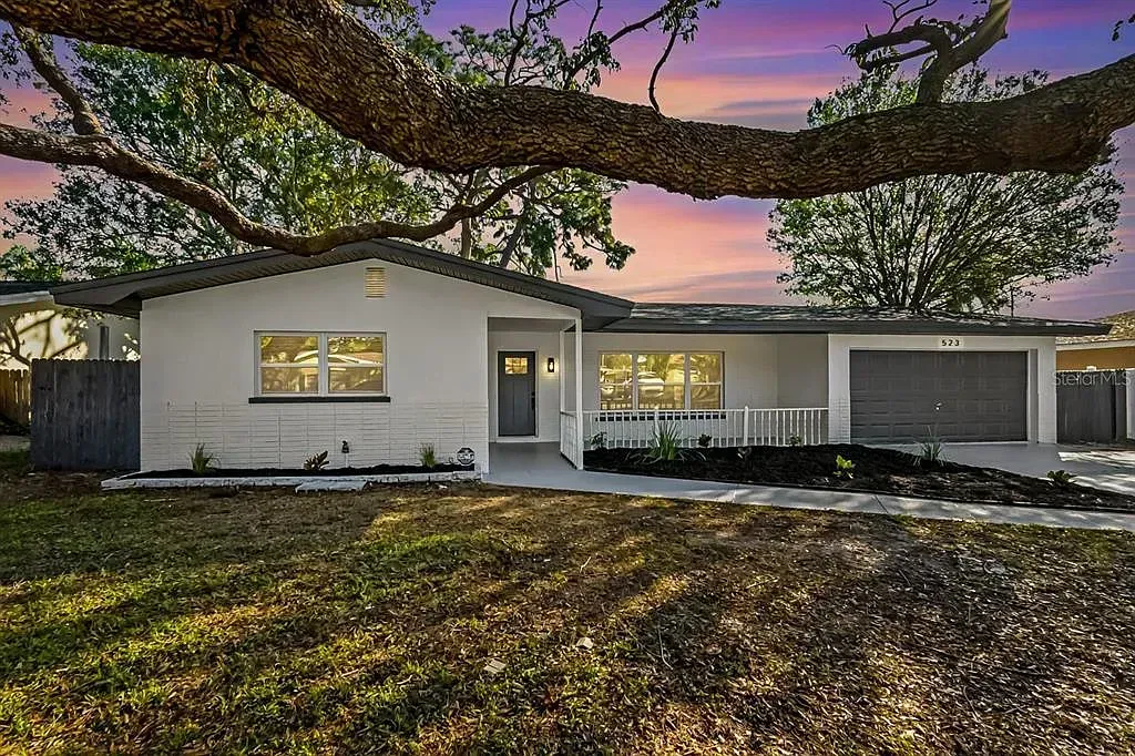 Single-story white house at dusk with lit windows, garage, and large tree branches overhead.