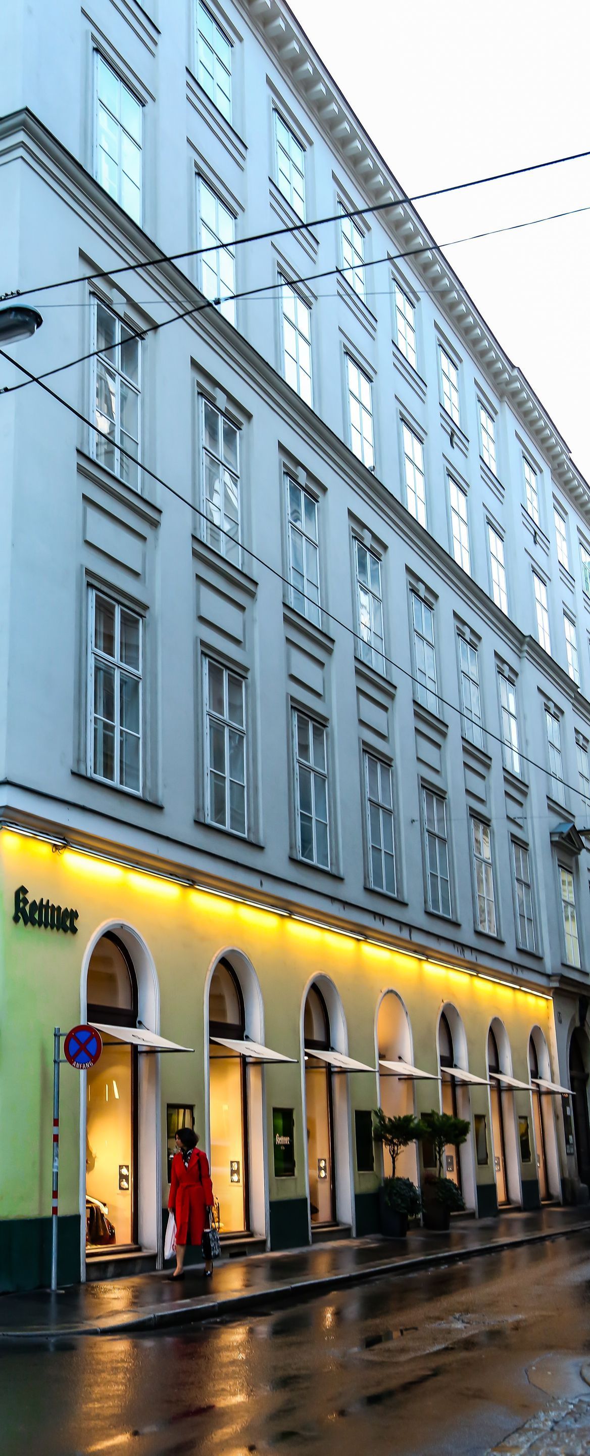 Rainy street view of a light blue building with yellow-lit arched storefronts and wet pavement