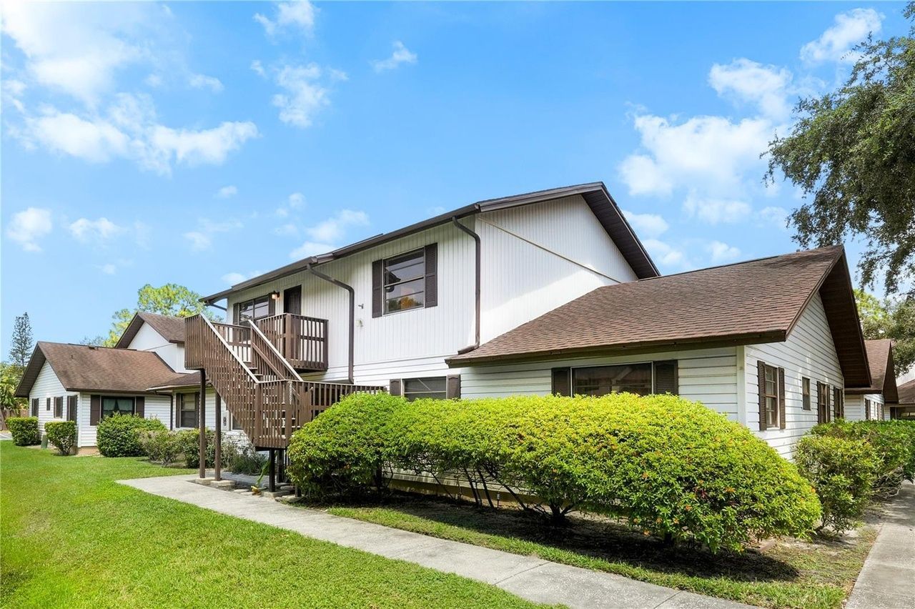 Two-story white apartment building with brown roof, stairs, and green lawn under a blue sky