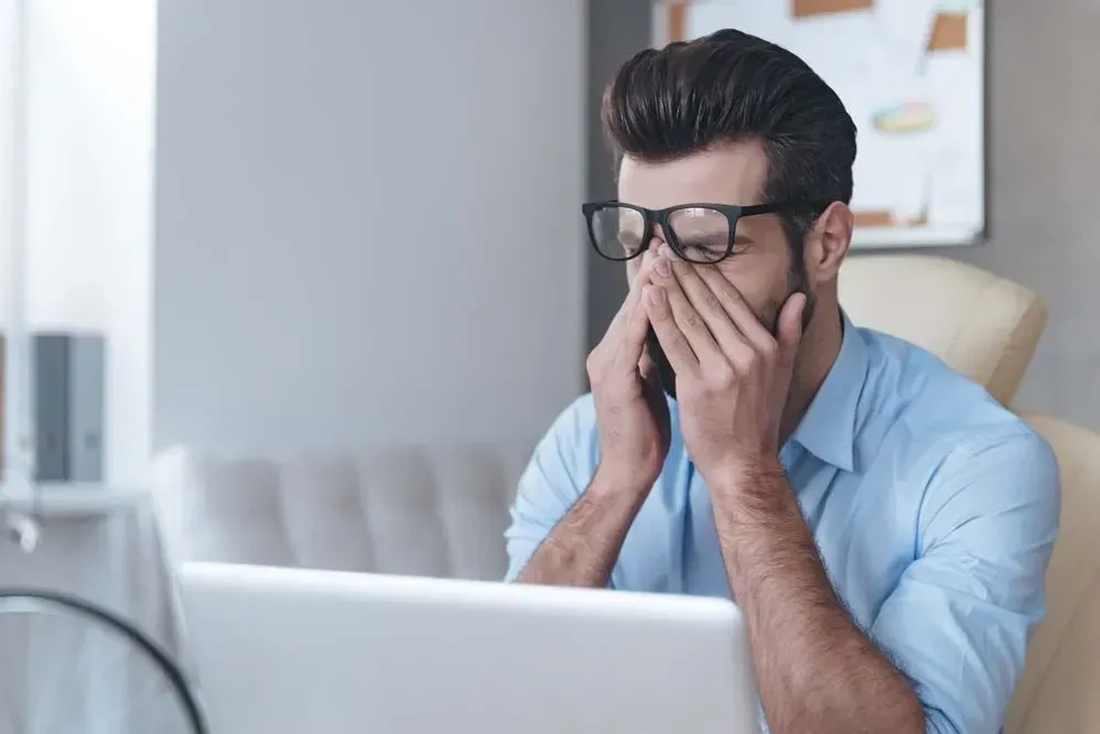 A man is sitting in front of a laptop computer covering his face with his hands.
