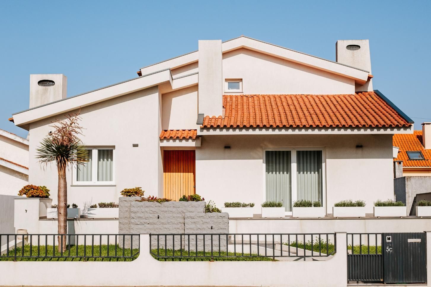 Modern white house with orange tile roof, front yard fence, and large windows under a clear sky