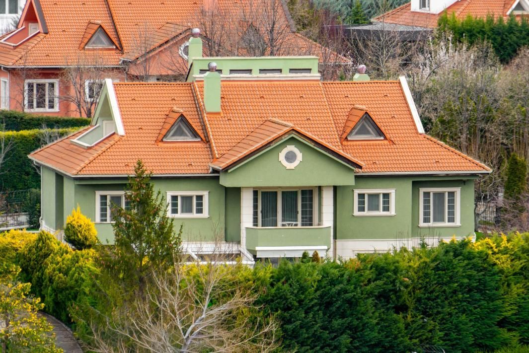 Green house with orange-tiled roof and white trim, surrounded by trees and hedges.