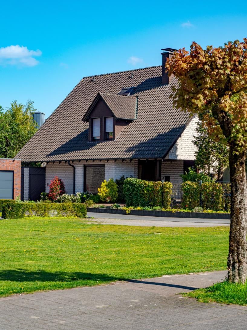 Suburban house with brown roof, green lawn, and a tree in the foreground under a blue sky