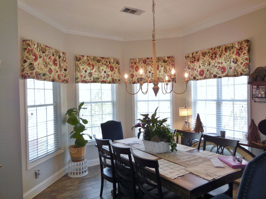 Dining room with large windows, floral valances, chandelier, and dark table and chairs.