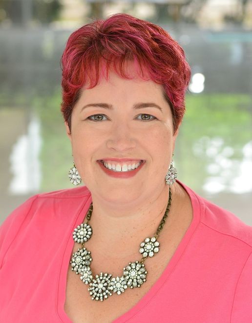 Smiling woman with short red hair wearing a pink top and silver floral necklace outdoors