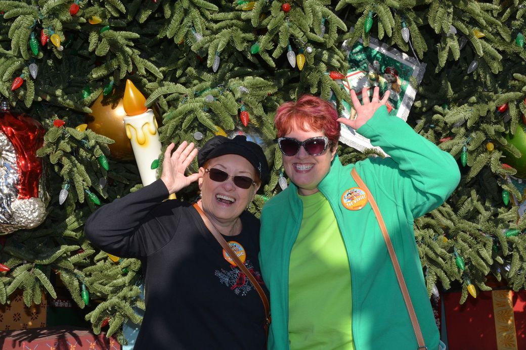 Two people in colorful outfits smiling and waving in front of a decorated Christmas tree