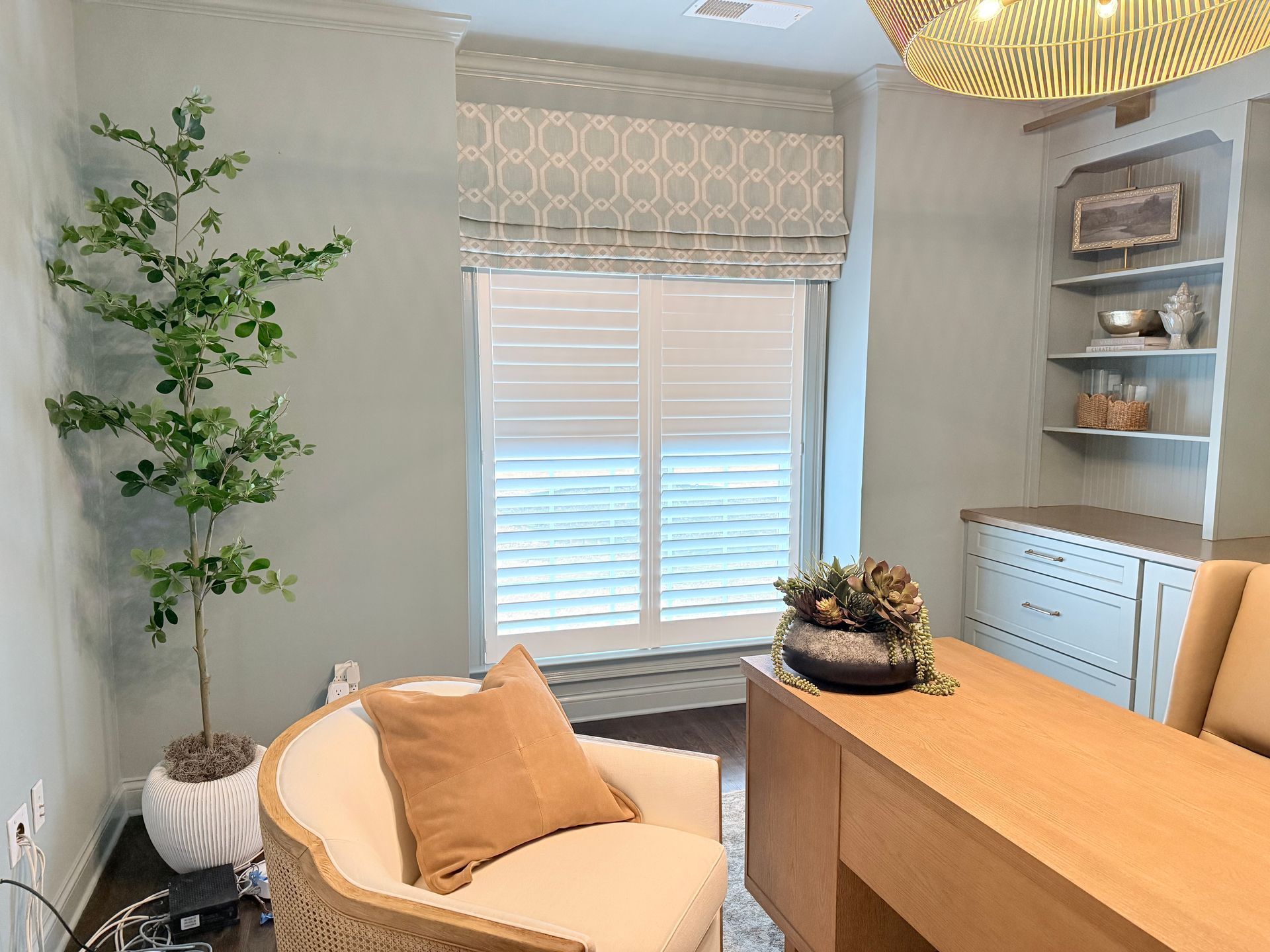 Modern home office with cream chair, wooden desk, window blinds, tall plant, and built-in shelves