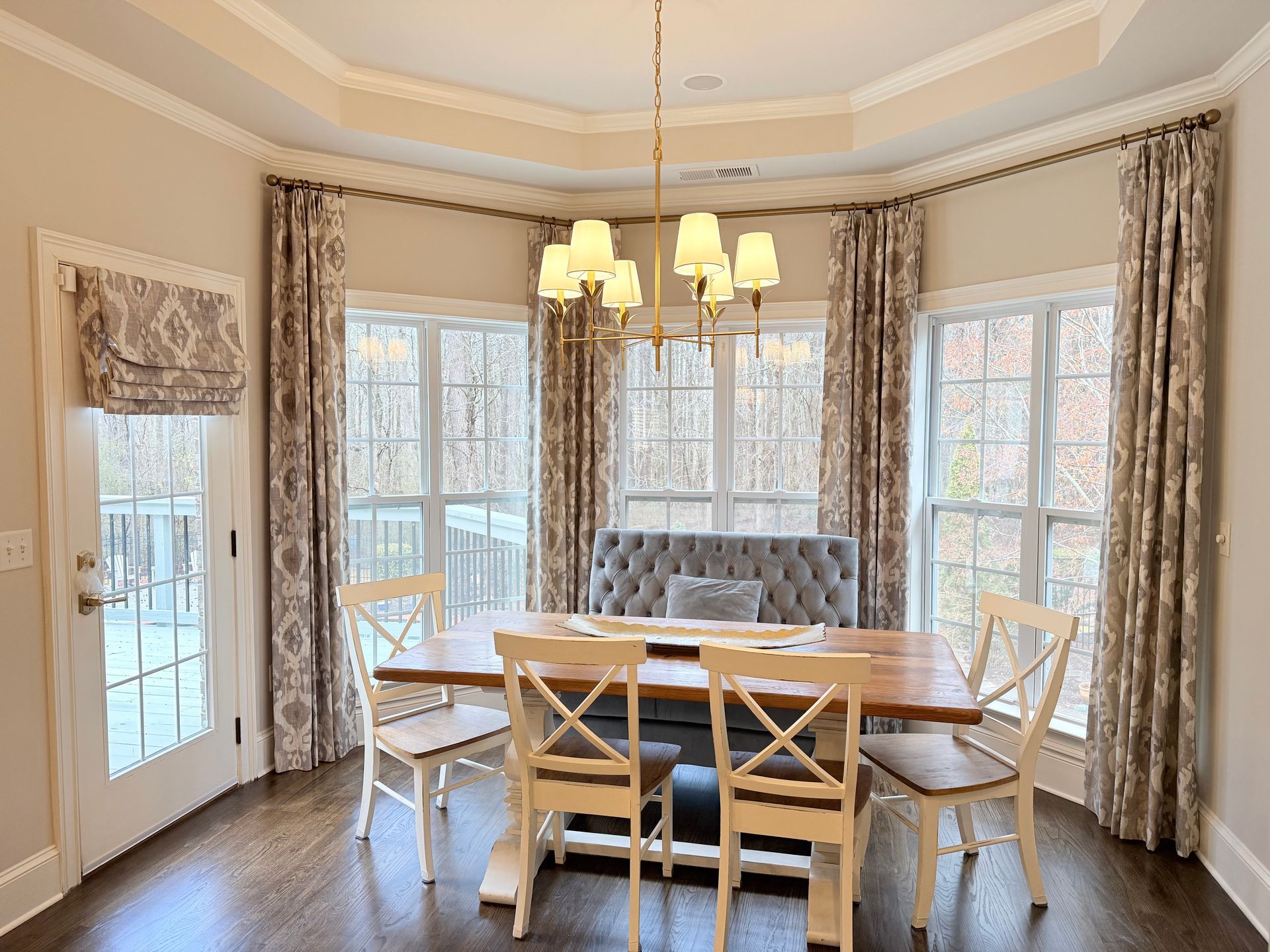 Bright dining room with round table, upholstered chairs, chandelier, and large windows with curtains