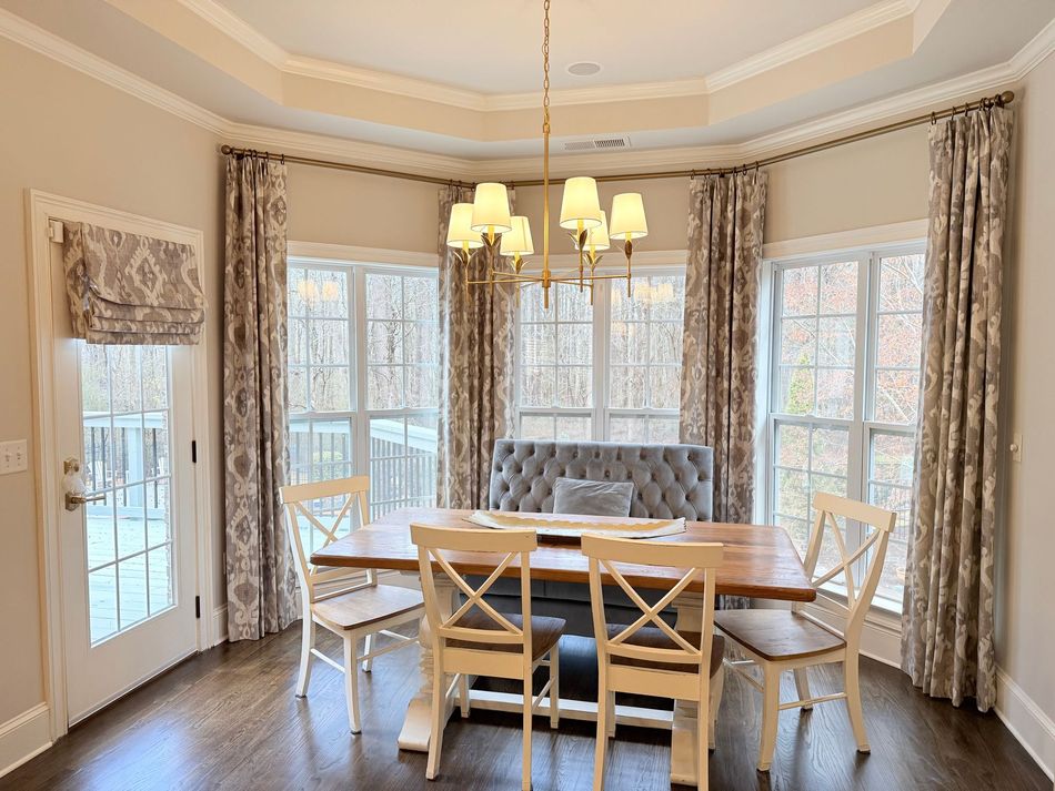Bright dining room with a round table, chairs, chandelier, and large windows with curtains