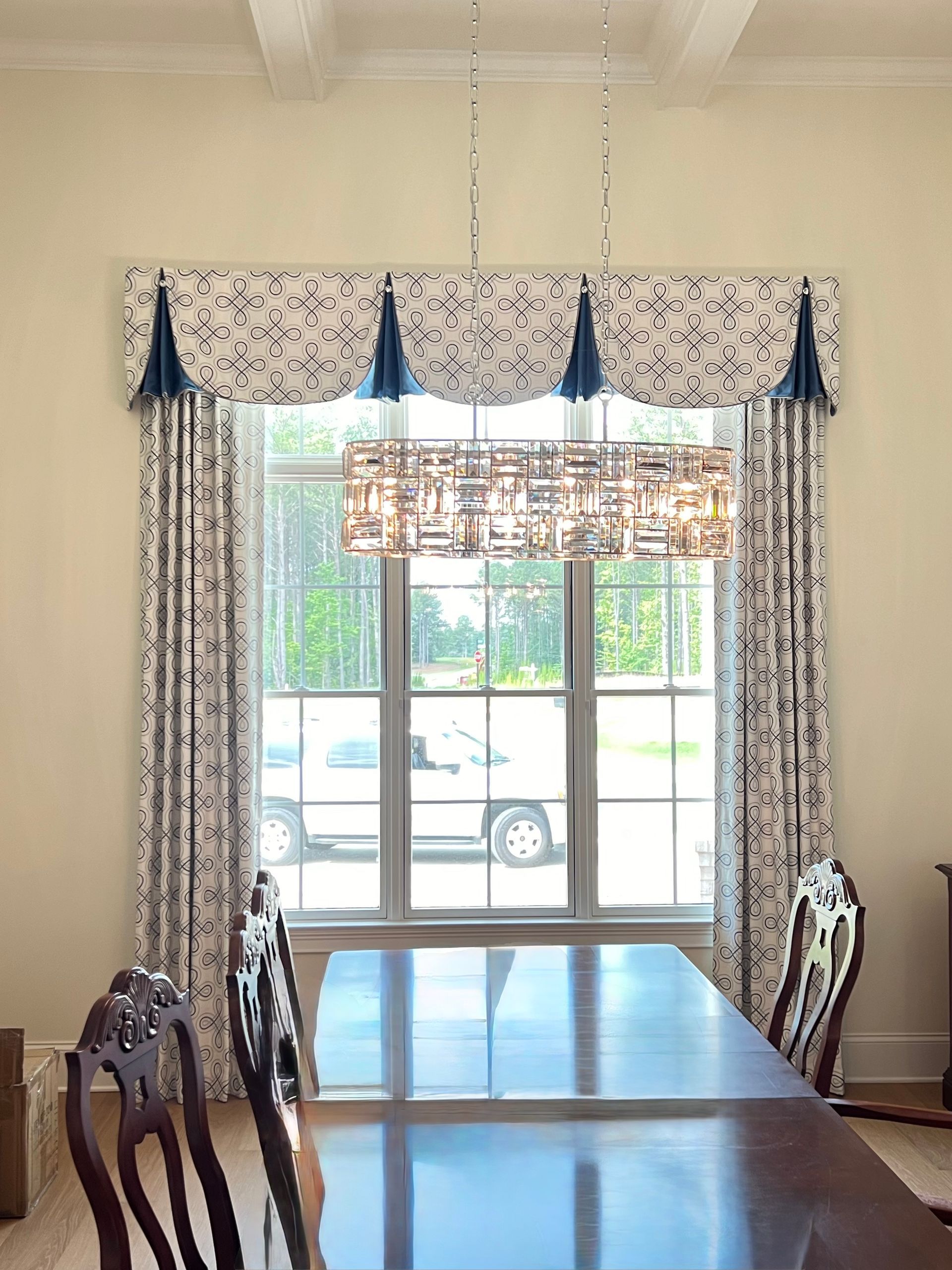 Dining room with a long table, patterned chairs, a chandelier, and large window drapes.