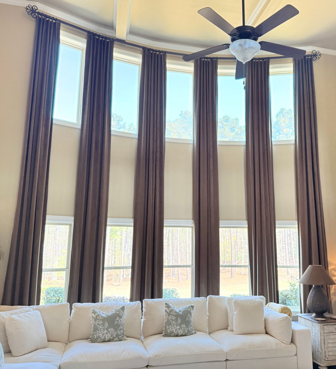Bright living room with tall windows, dark curtains, a white sofa, and a ceiling fan.