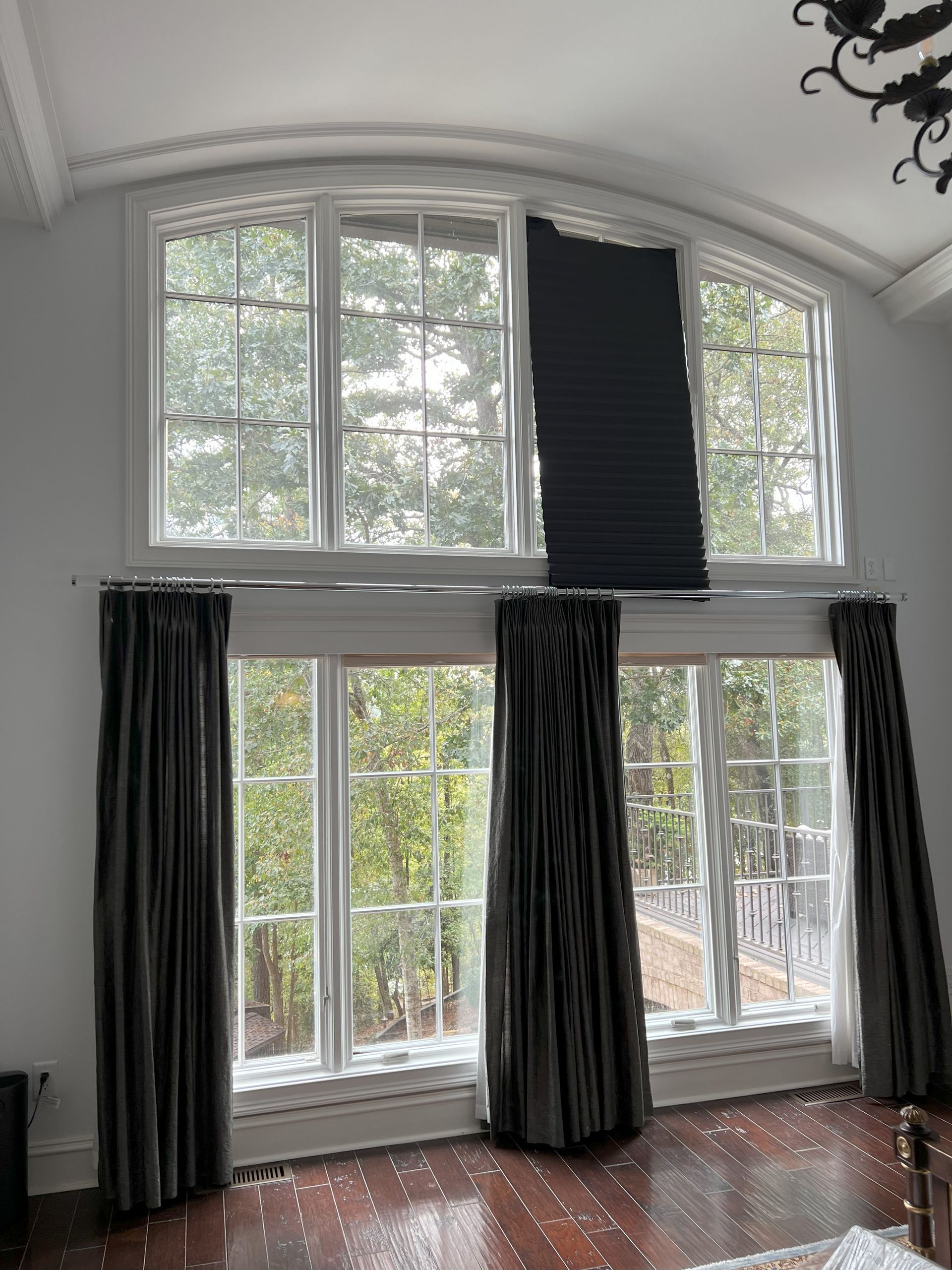Tall white living room windows with dark curtains and a black blind, showing trees outside.