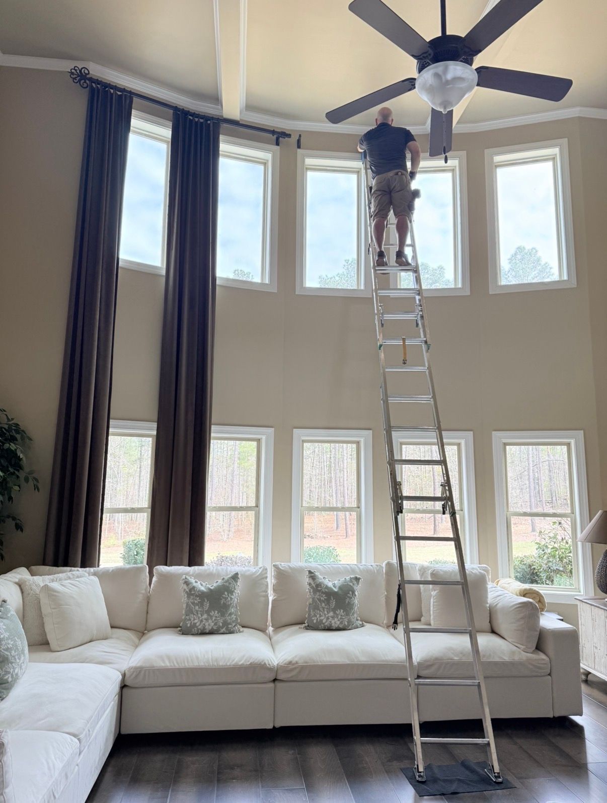Person on a ladder adjusting a ceiling fan in a bright living room with tall windows and a white sectional sofa