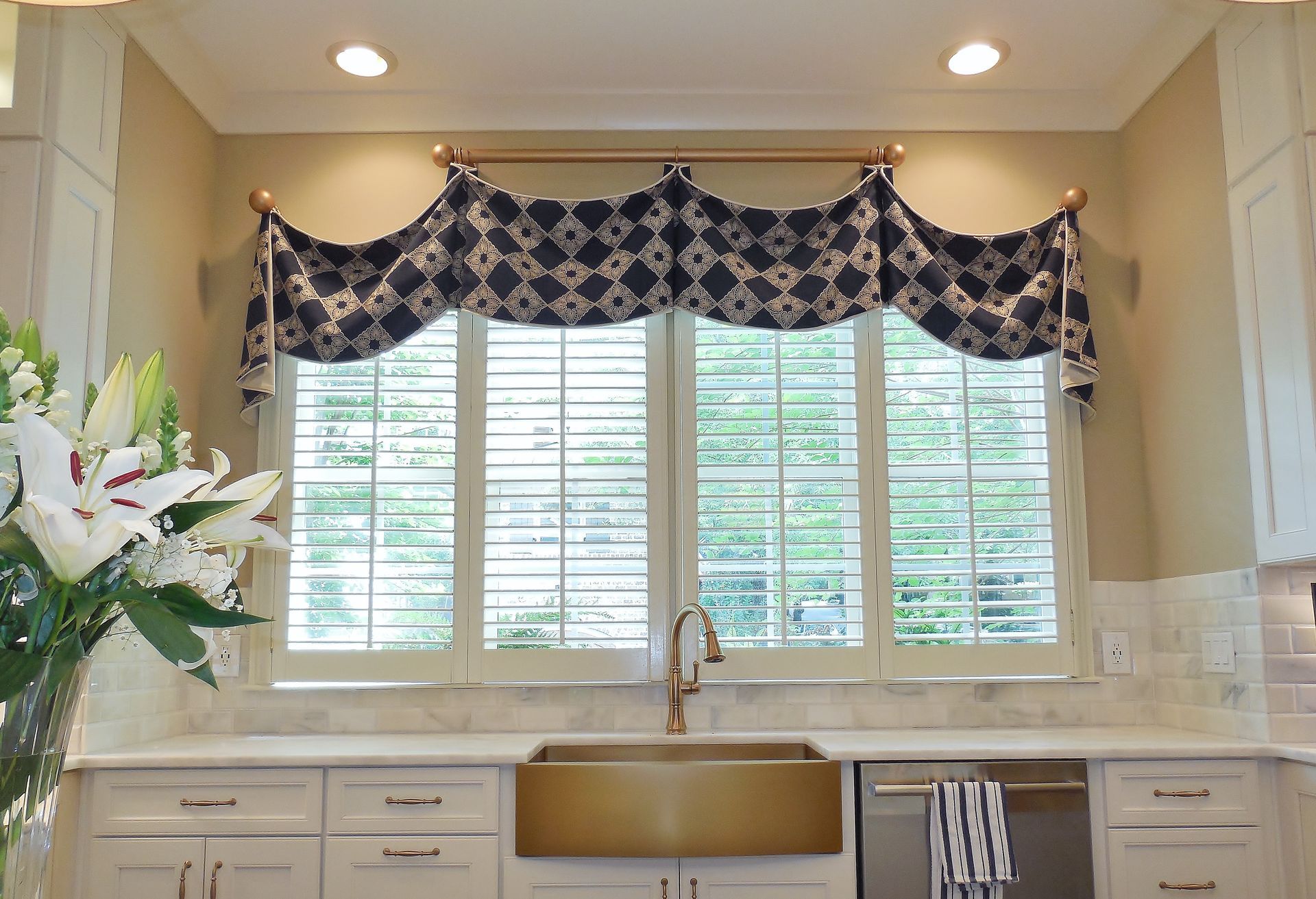 Bright kitchen window with patterned valance, white cabinets, gold sink, and a vase of white flowers.
