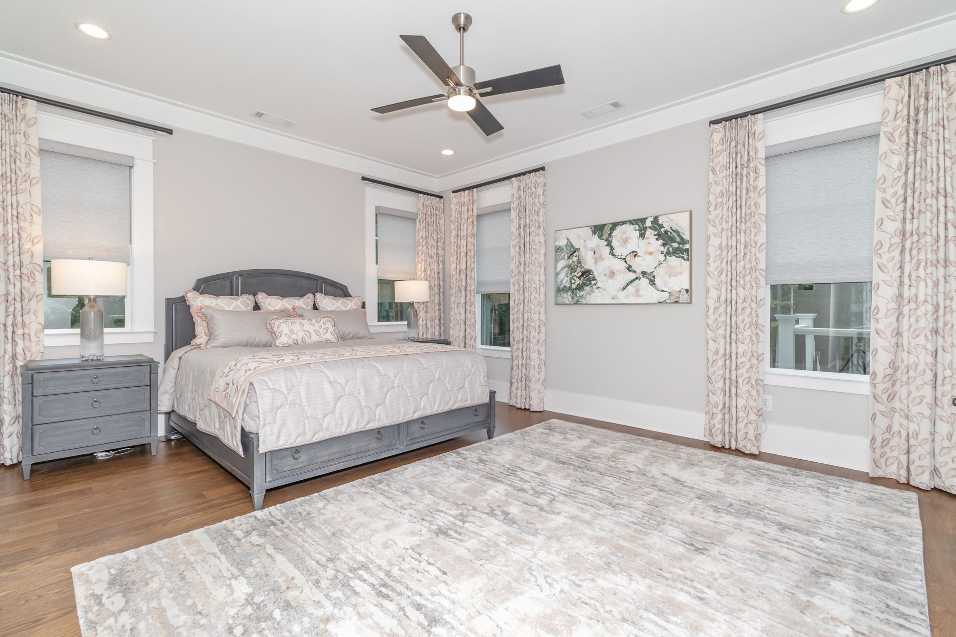 Spacious gray bedroom with a bed, nightstands, large rug, ceiling fan, and tall windows with curtains