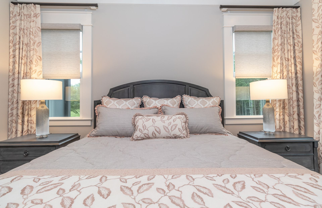 Bedroom with a gray upholstered bed, patterned bedding, bedside lamps, and two windows with curtains
