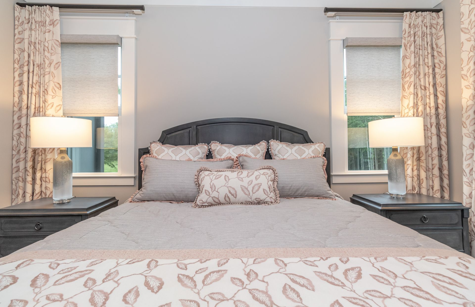 Bedroom with a gray upholstered bed, patterned bedding, bedside lamps, and two windows with curtains