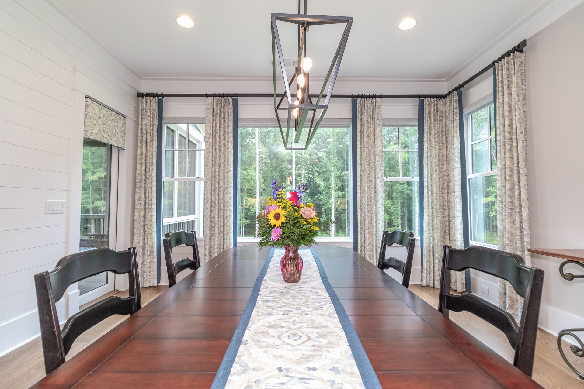 Bright dining room with long wooden table, black chairs, floral centerpiece, and large windows overlooking greenery