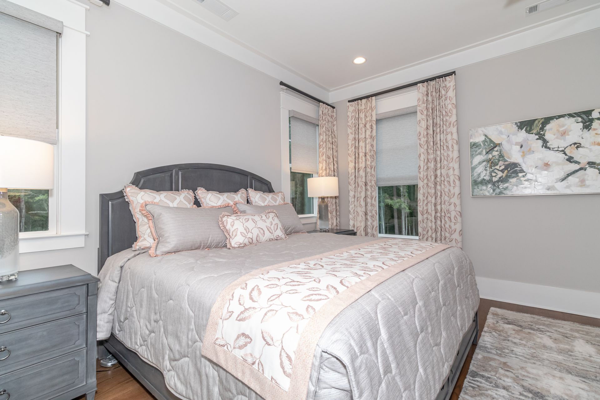 Bright bedroom with a gray upholstered bed, patterned bedding, nightstand, and floral wall art