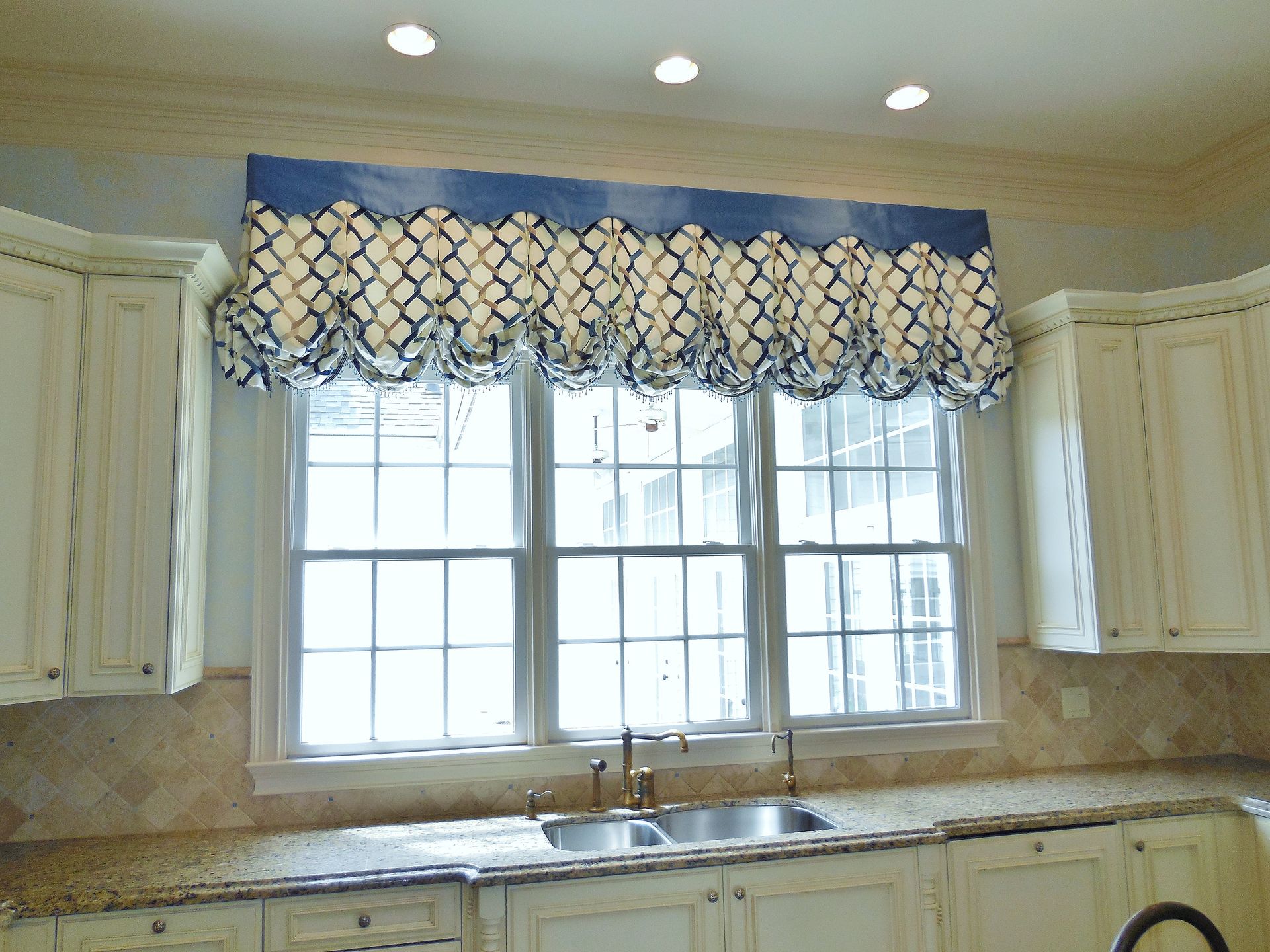 Kitchen with cream cabinets, blue patterned valance, and a double window over a countertop sink.