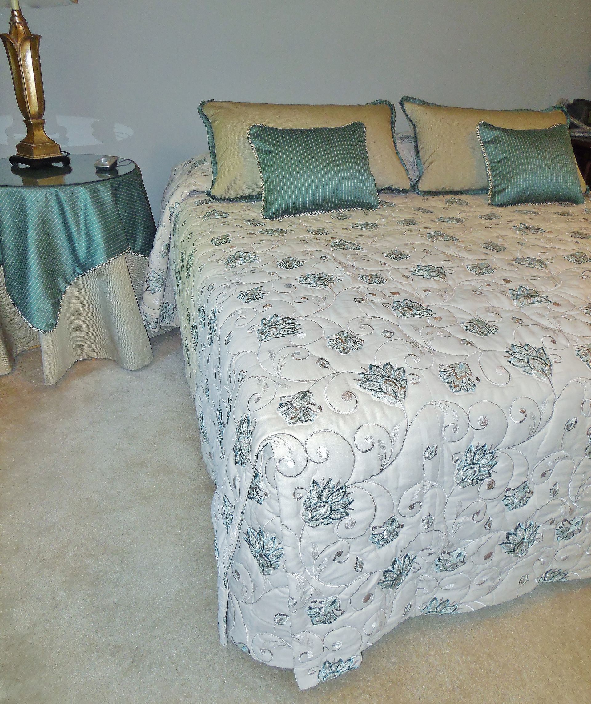 A bedroom with a bed dressed in white floral bedding and green pillows beside a small table.
