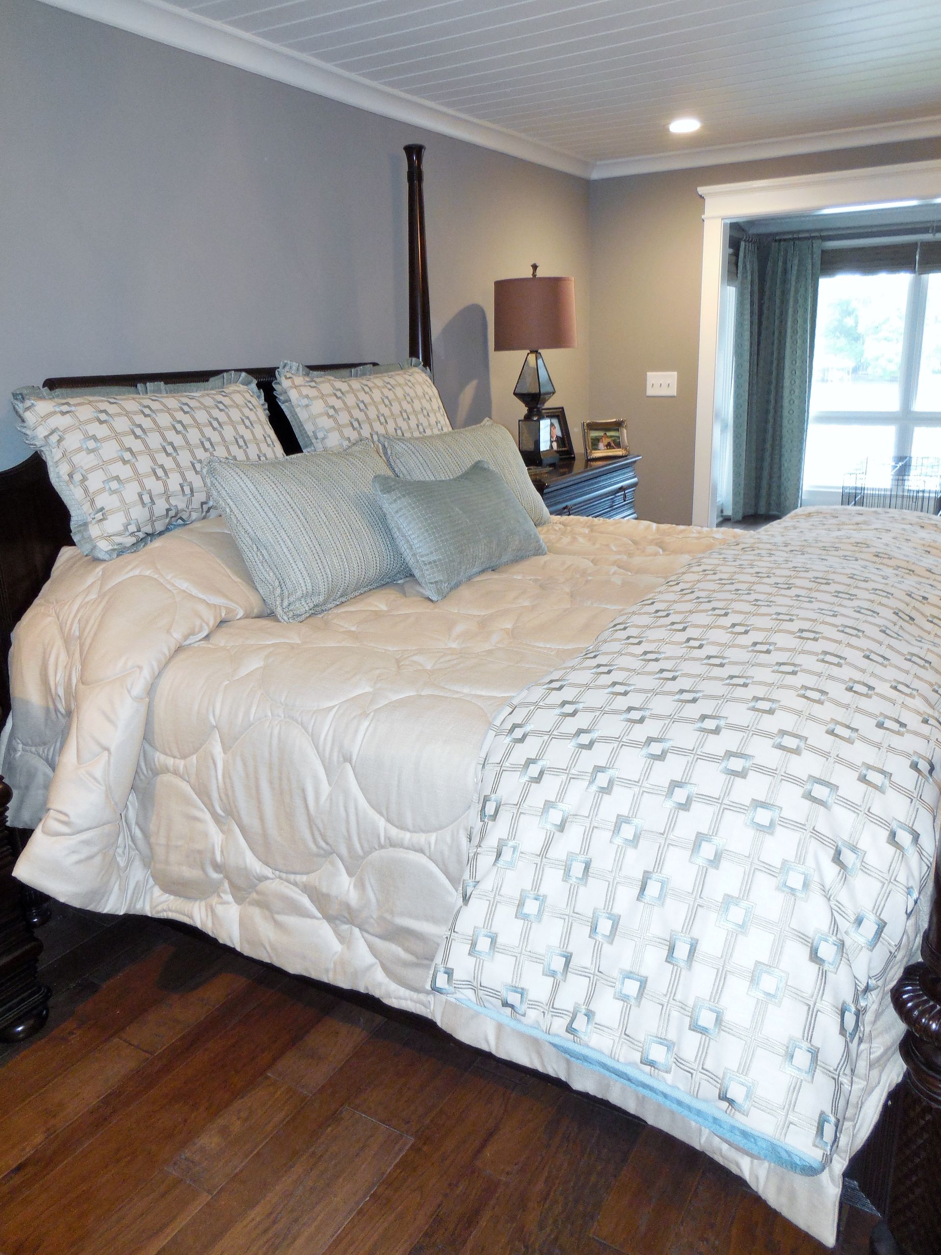 Bedroom with white quilted bed, blue patterned pillows, wooden floor, and lamp by a window.