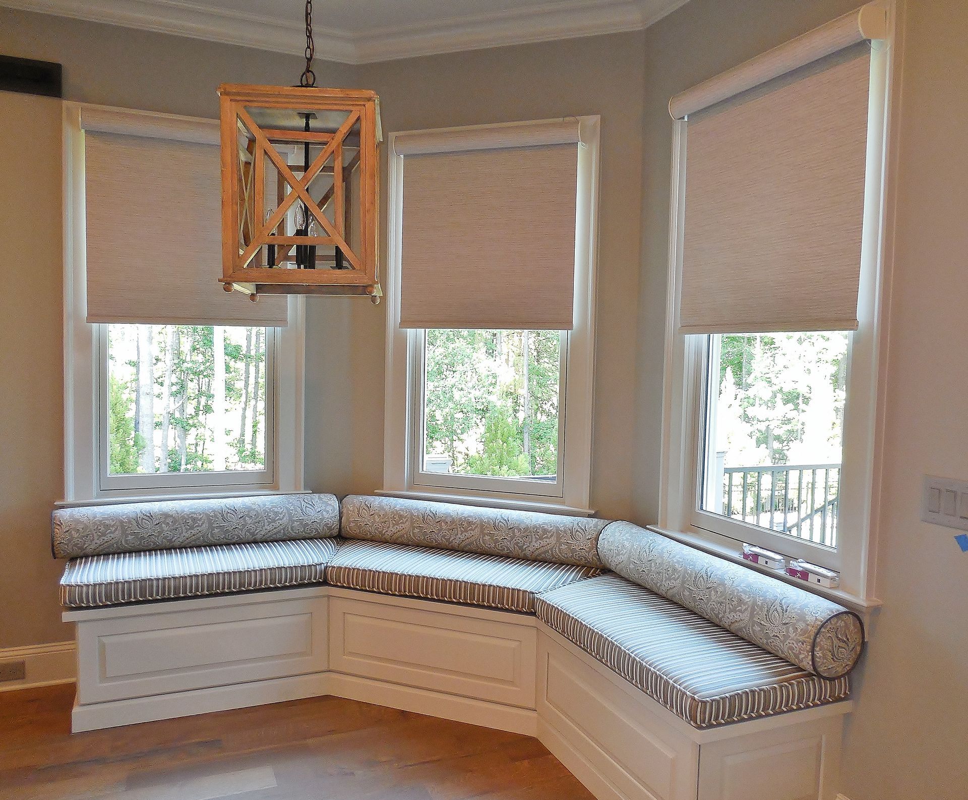 Curved window seat with white drawers, patterned cushion, and three beige shades in a bright corner room