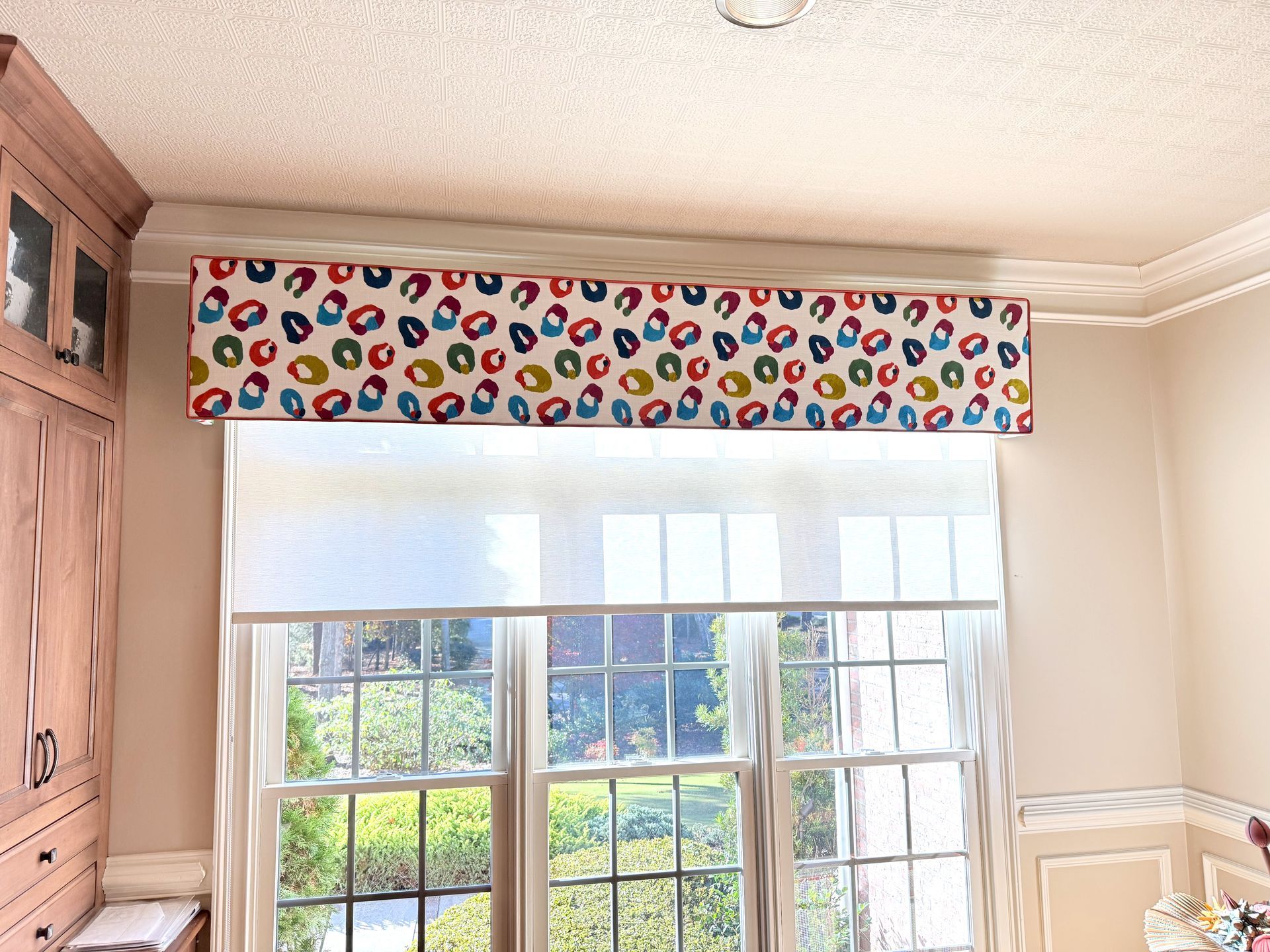 Bright bedroom window with colorful patterned valance and white blinds, sunlight and garden view outside