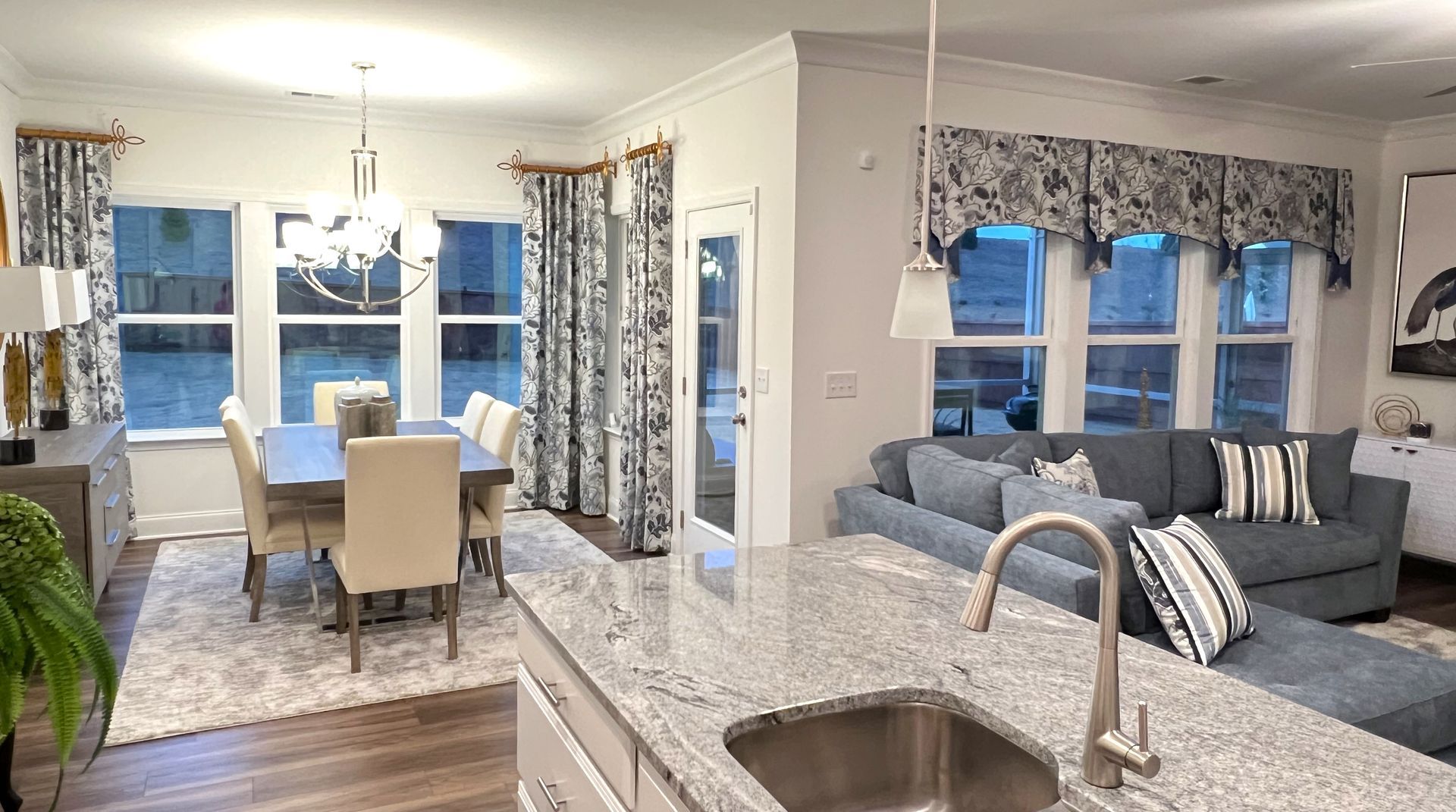 Open-plan kitchen and dining area with granite island, gray sofa, and large windows with patterned curtains