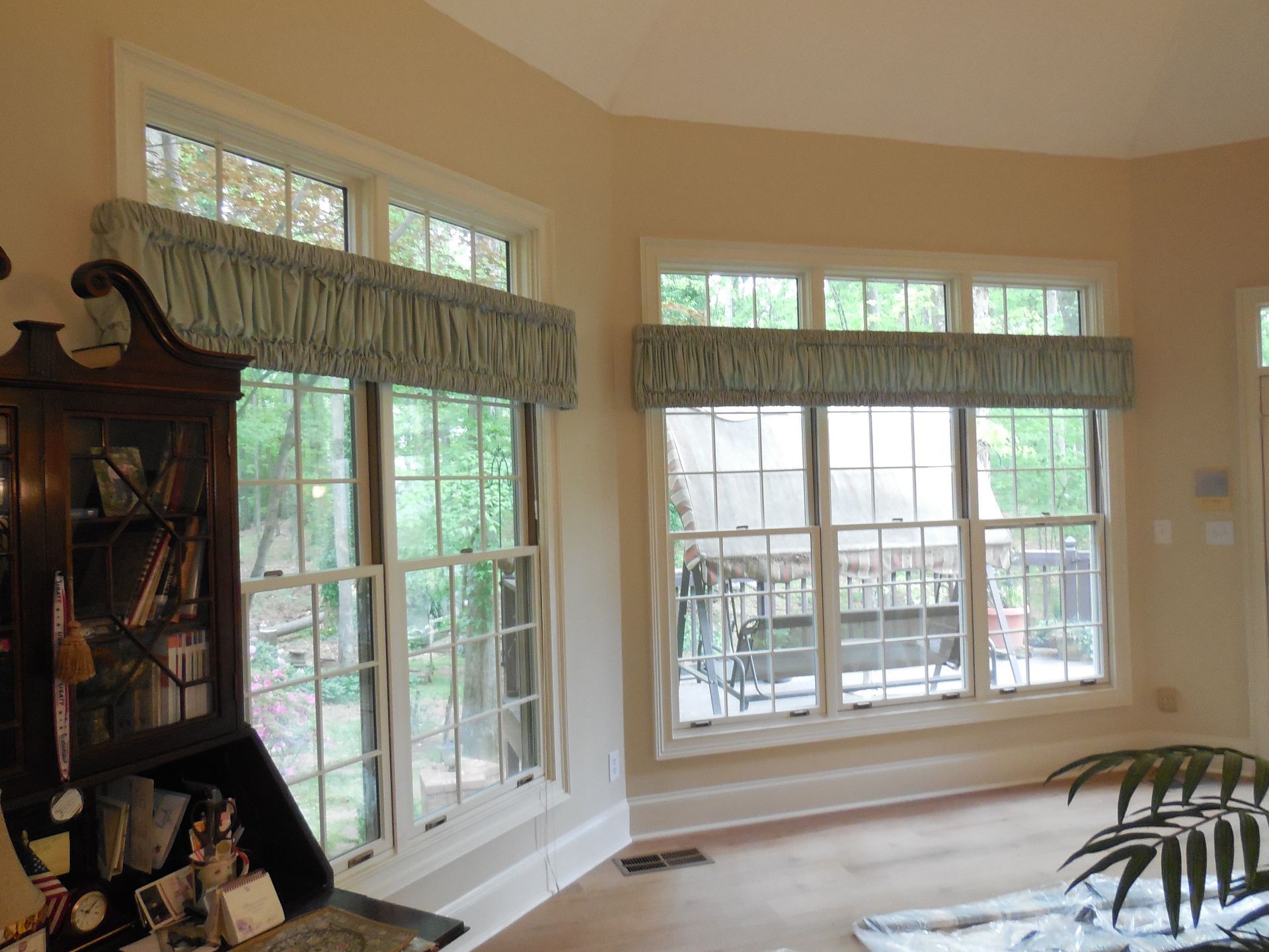 Bright living room with large windows, blue-green valances, and a dark wooden cabinet.