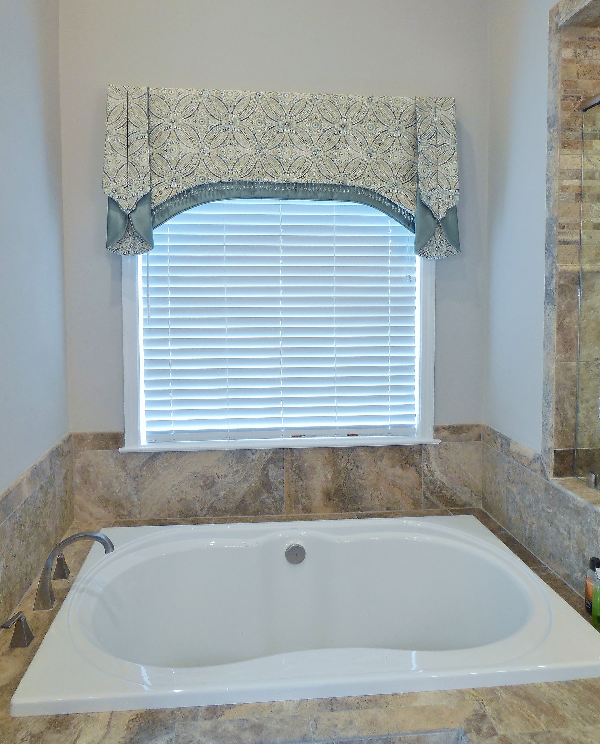 Bathroom with a white soaking tub beneath a window with blinds and floral valance