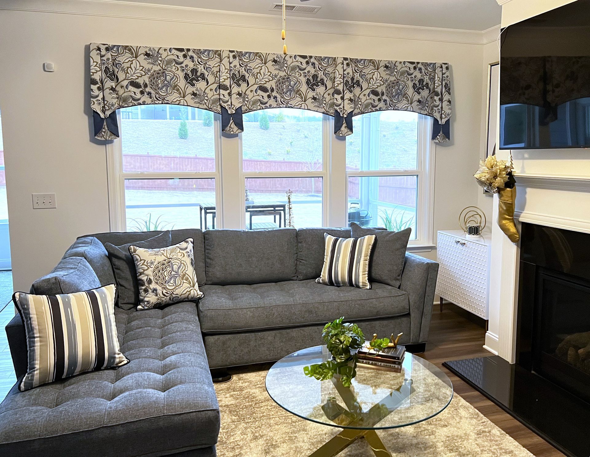 Gray sectional sofa in a bright living room with patterned window valance, glass coffee table, and fireplace.