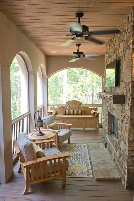 Covered porch with wooden seating, stone fireplace, arched openings, and ceiling fans overlooking greenery