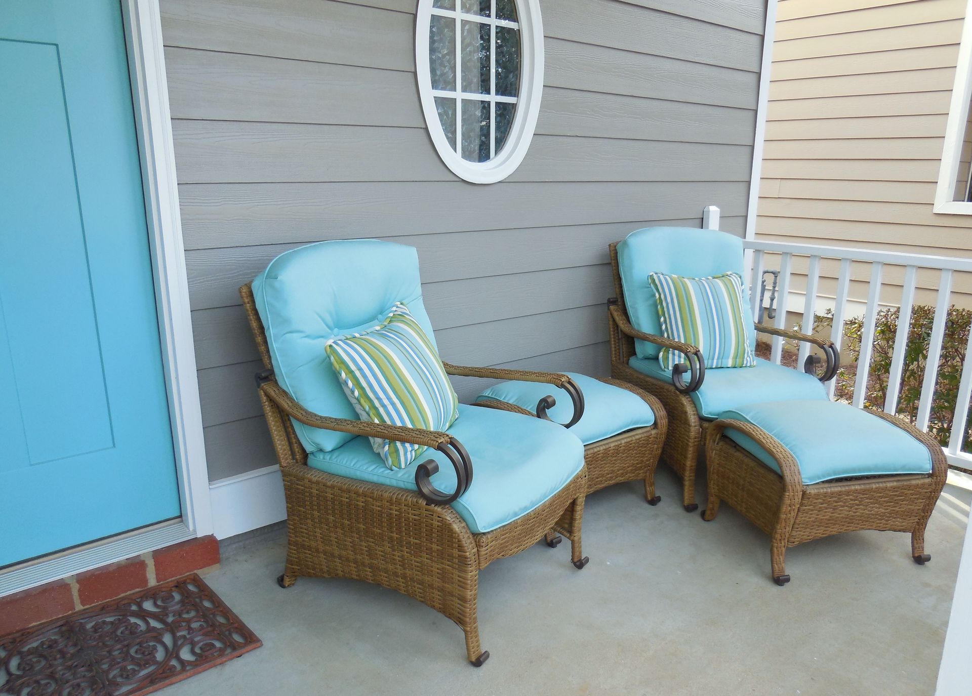 Two turquoise cushioned wicker chairs on a porch beside a blue door and white railing.