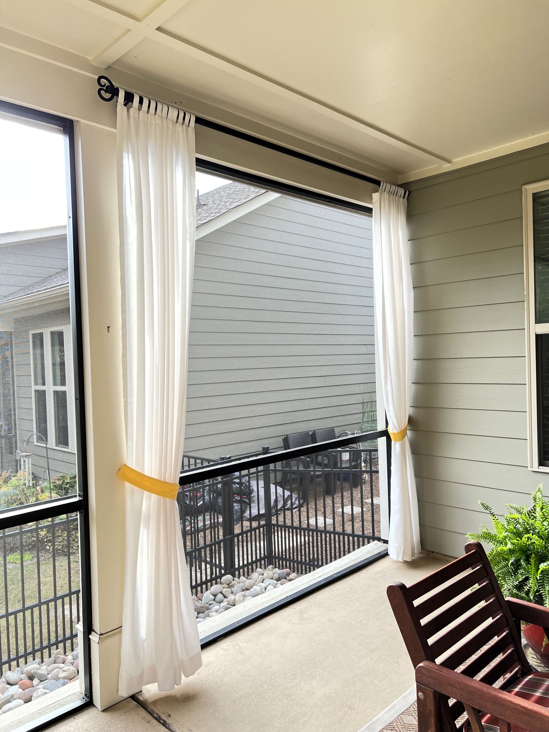 Covered balcony with white curtains, black railing, wooden chair, and potted plants overlooking neighboring houses