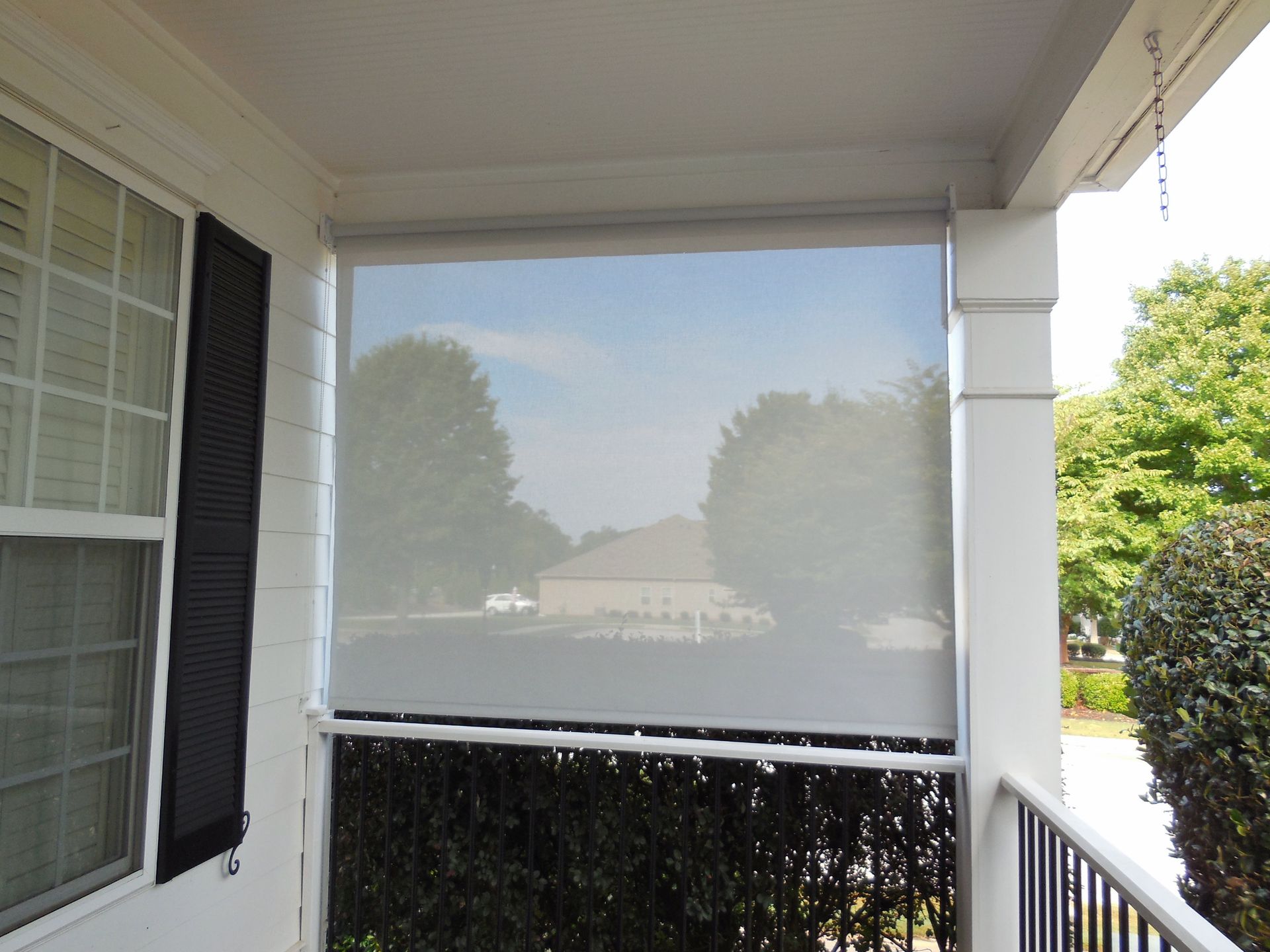 Front porch with a white mesh shade screen and black shutters, overlooking a sunny yard.