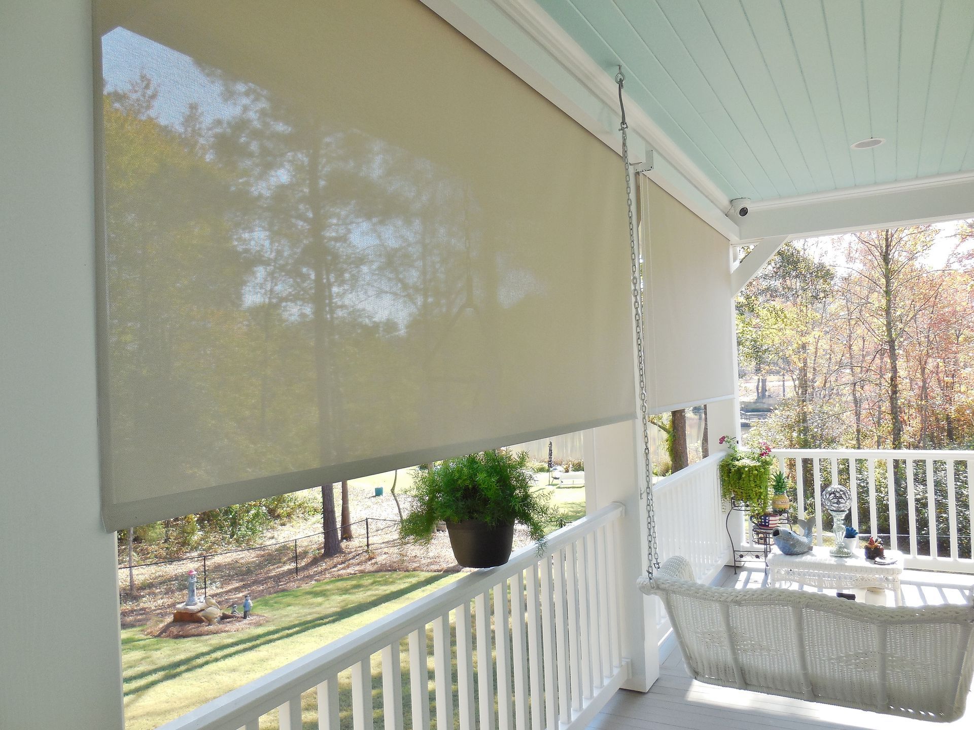 Covered porch with white railing, beige roll-down shade, and potted plants overlooking a wooded yard