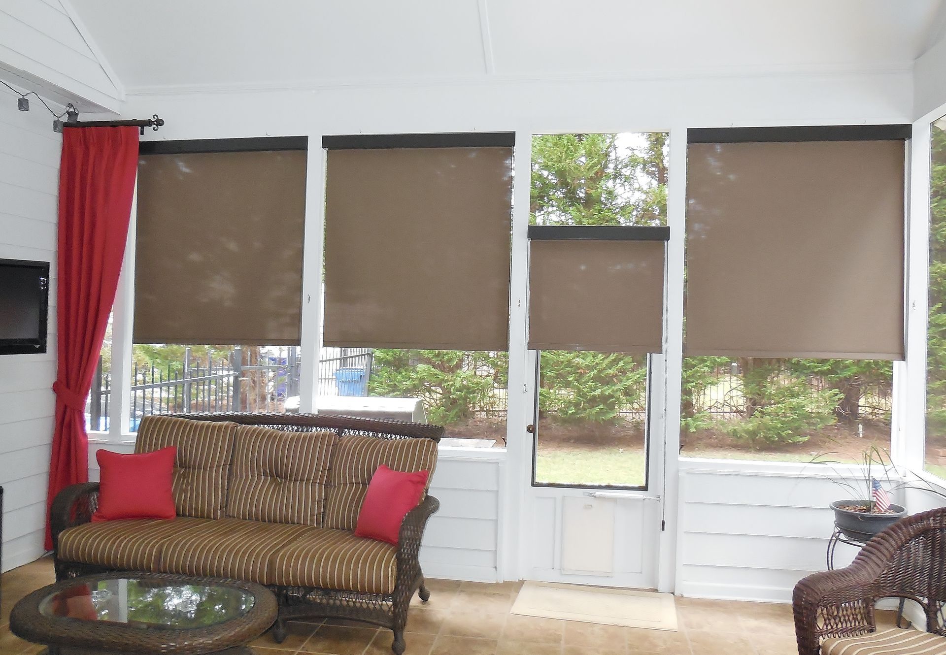 Sunroom with wicker furniture, red cushions, and beige window blinds overlooking trees.