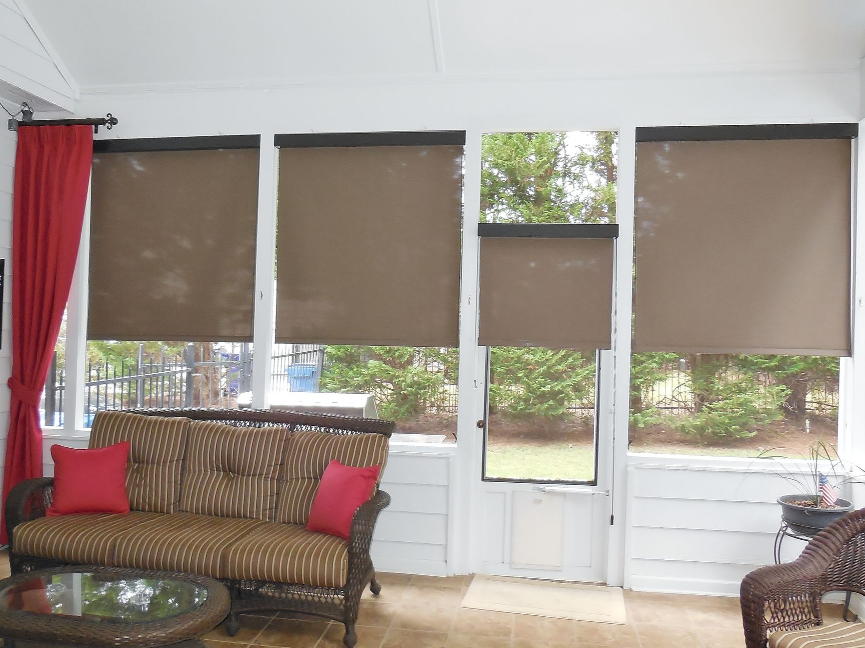 Sunroom with wicker sofas, red cushions, and beige shades over large windows and a glass door