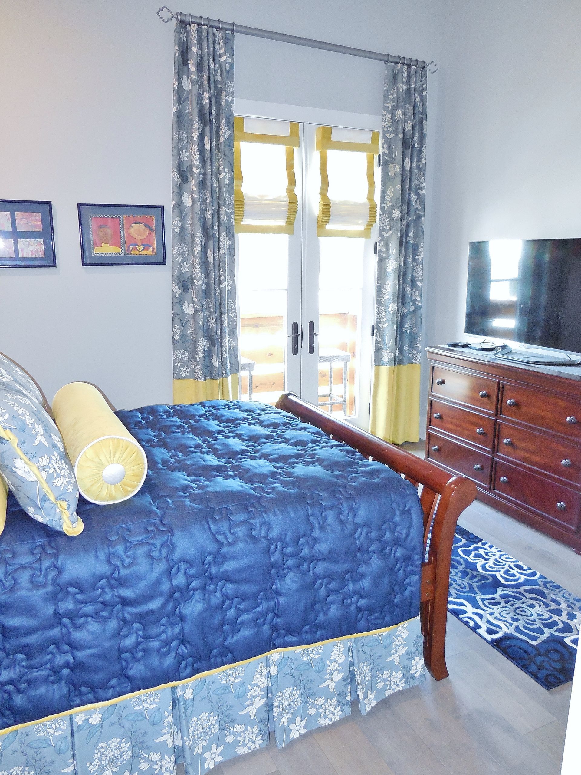 Blue bedroom with bed, dresser, TV, and window with patterned curtains
