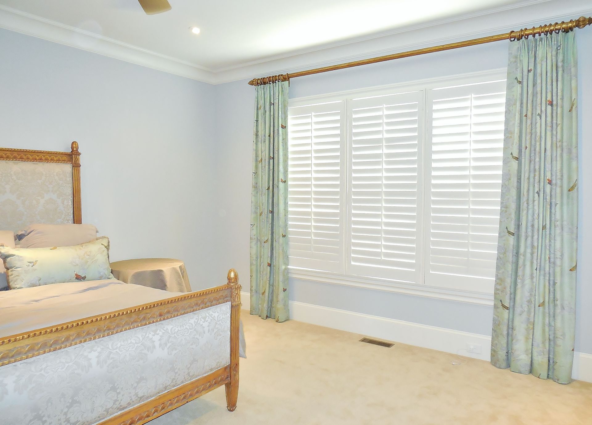 Bright bedroom with a wooden bed, cream carpet, and large window with pale blue curtains and white shutters