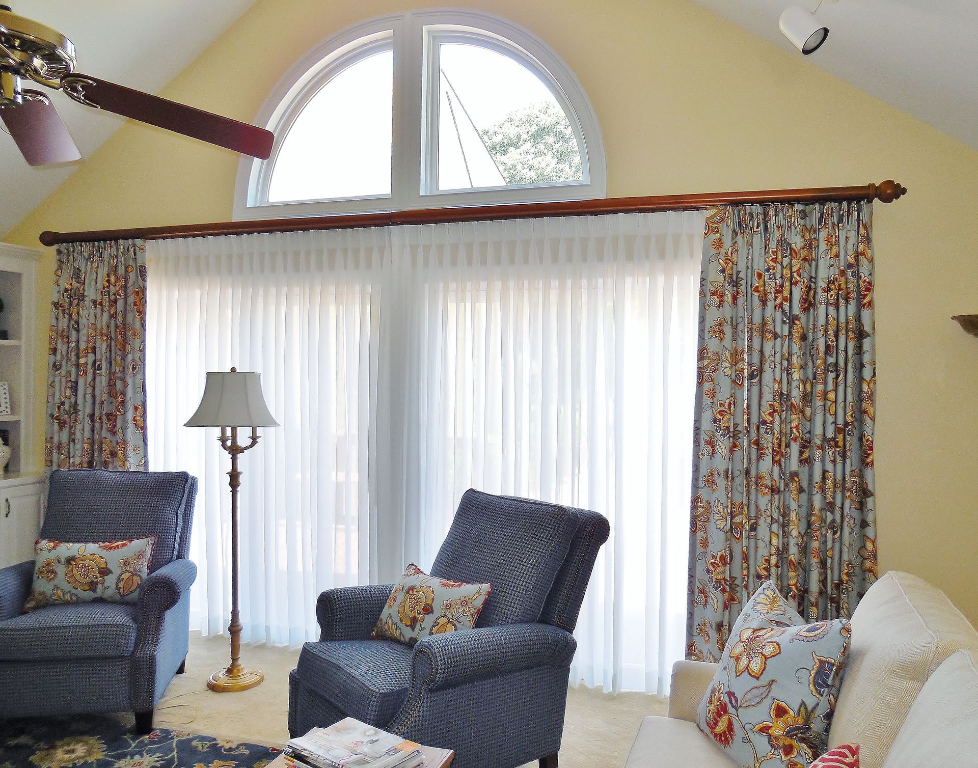 Bright living room with blue armchairs, floral curtains, floor lamp, and large window with sheer drapes
