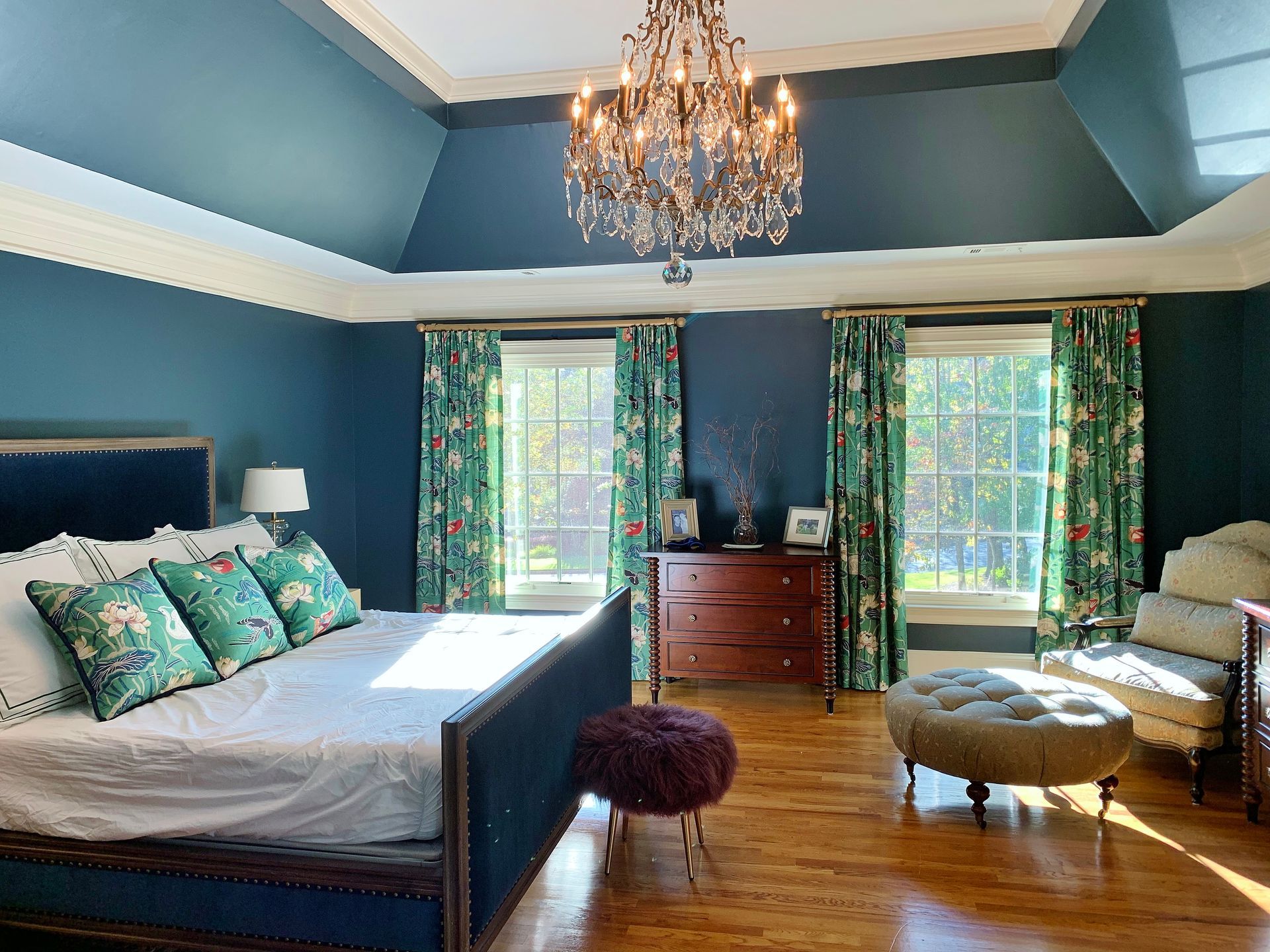 Blue bedroom with a dark wood bed, chandelier, patterned curtains, and a chaise lounge by the window.
