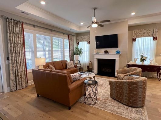Bright living room with brown sofa, wicker chairs, fireplace, TV, and large windows with curtains.