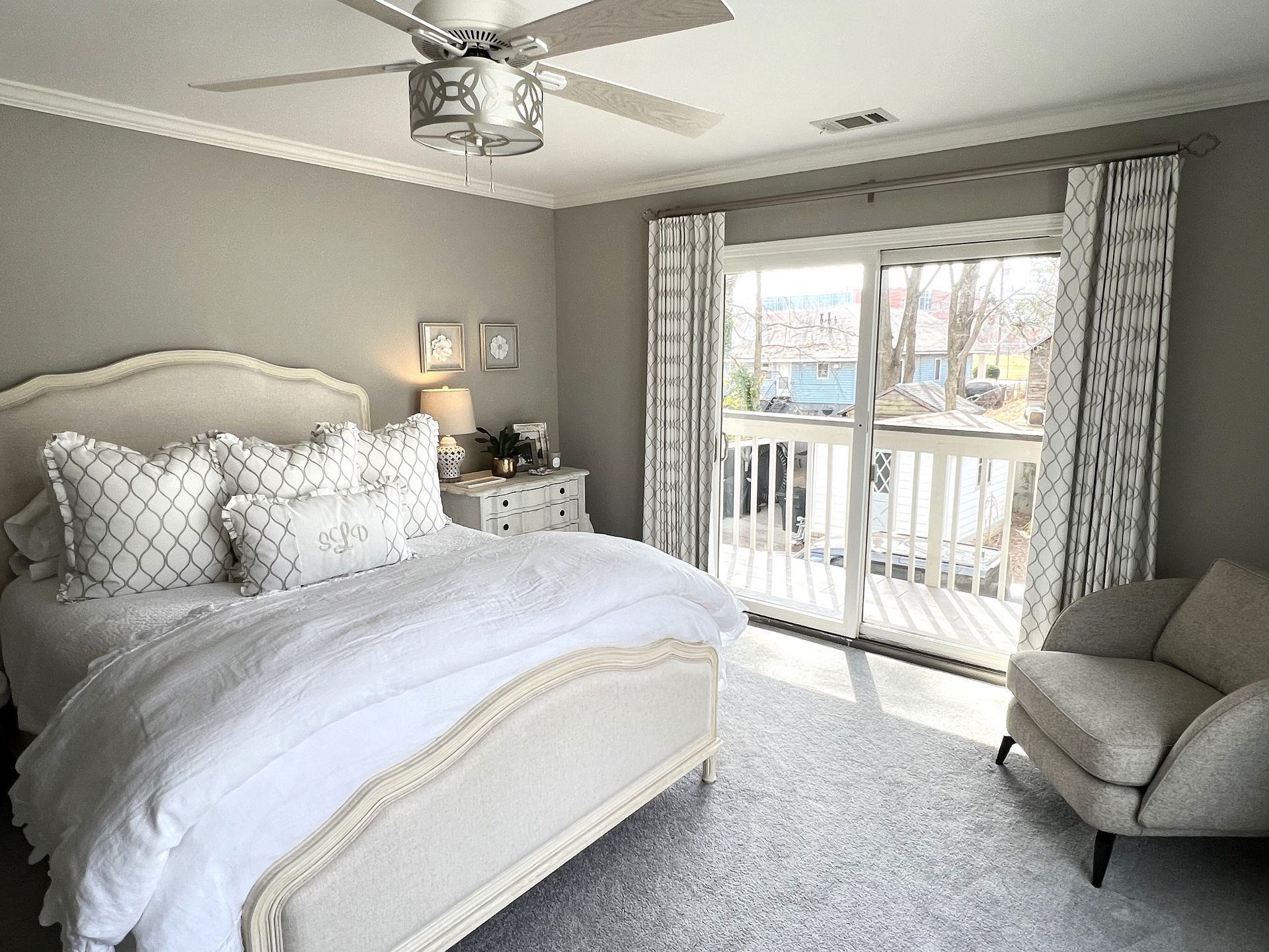 Bright bedroom with a white bed, gray armchair, ceiling fan, and balcony doors with curtains