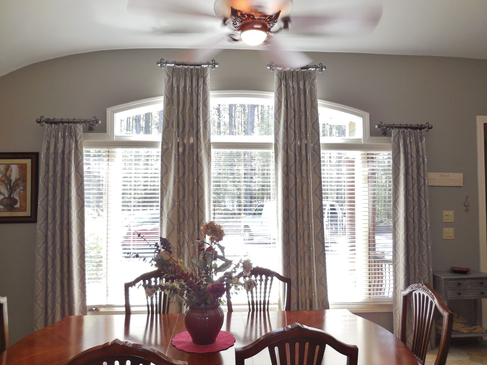 Dining room with large arched windows, curtains, and a table with a flower centerpiece beneath a ceiling fan.