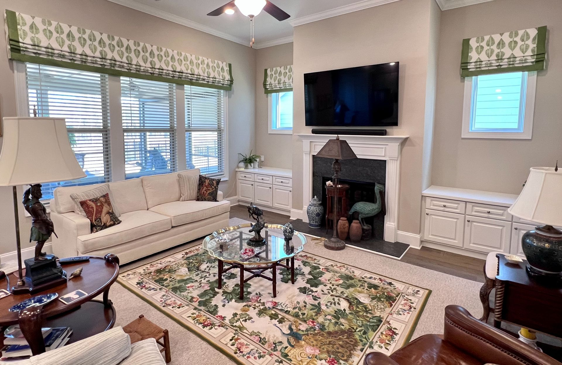 Elegant living room with beige sofa, floral rug, fireplace, wall-mounted TV, and large windows