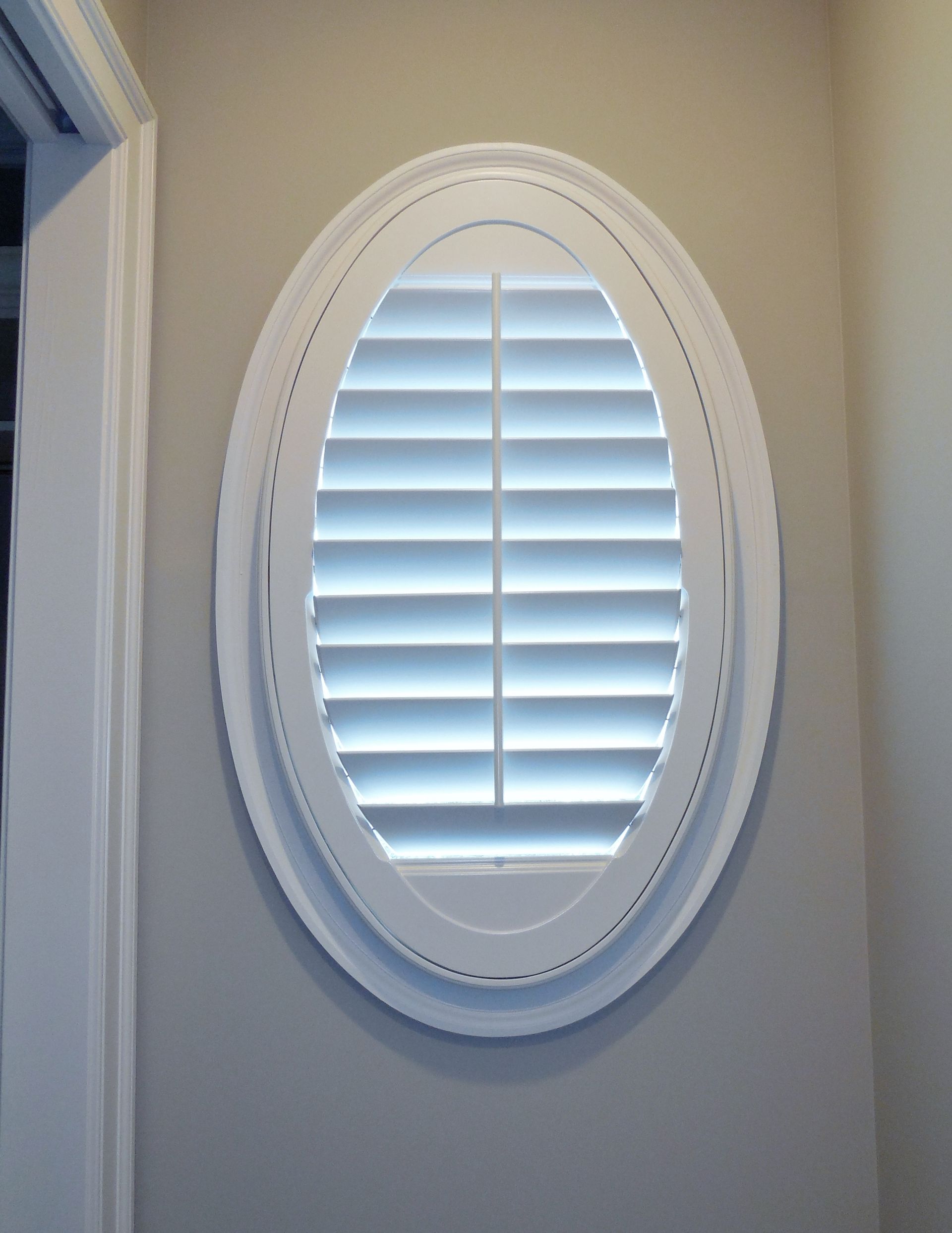Oval white window with closed white blinds on a light gray wall