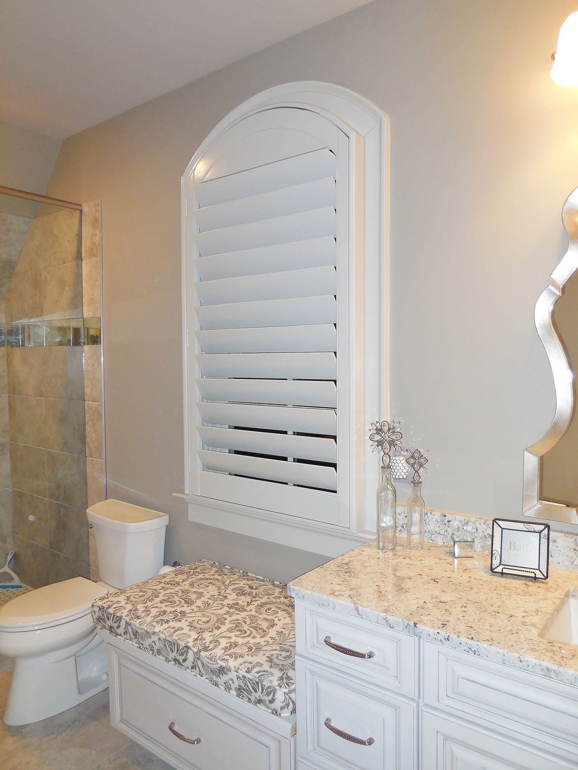 Elegant bathroom with white vanity, granite countertop, toilet, and arched shuttered window.